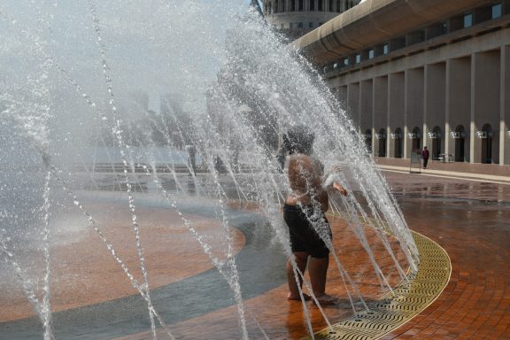 Child playing in water fountain on a sunny day, surrounded by a plaza.