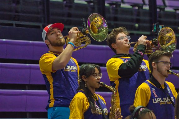 Band members play brass instruments in a gymnasium, wearing matching uniforms.