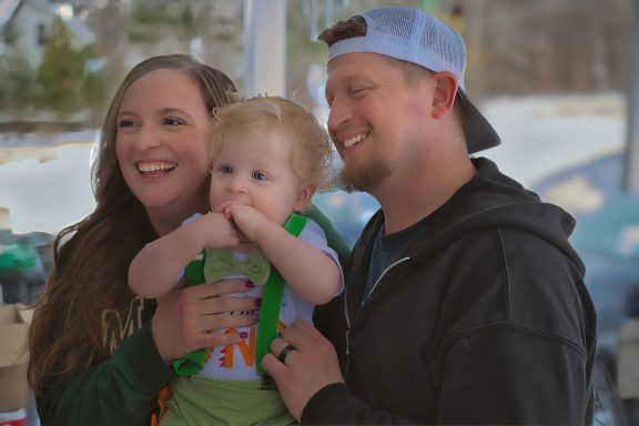A smiling family poses outdoors with a toddler in the mother's arms.