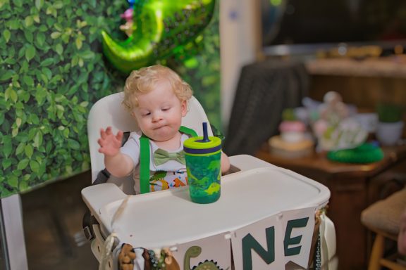 Toddler in a high chair waves, holding a green cup, with festive decorations in the background.