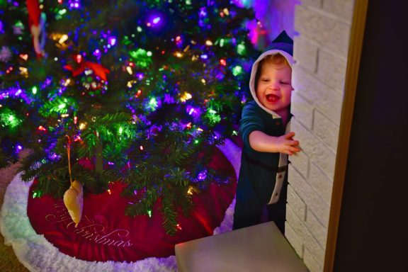 Child next to Christmas Tree Child in a hooded outfit smiling next to a brightly lit Christmas tree.