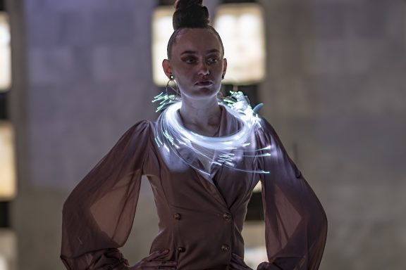 A model poses in a stylish, illuminated outfit with a unique necklace.