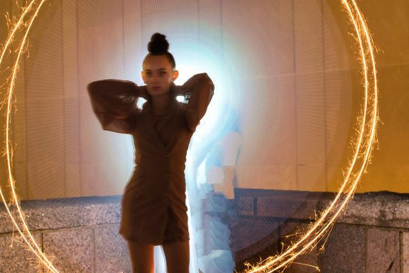 A person in a brown outfit poses with a glowing circular light behind them.