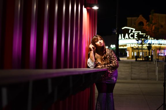 A woman leans against a purple-lit barrier at night, with colorful city lights in the background.